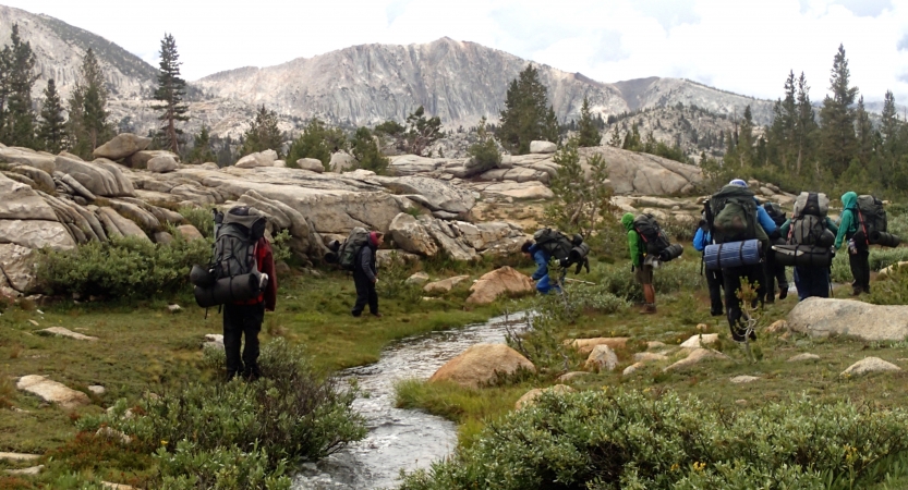 A group of outward bound students wearing backpacks hike beside a stream in a grassy field. There are mountains in the distance.